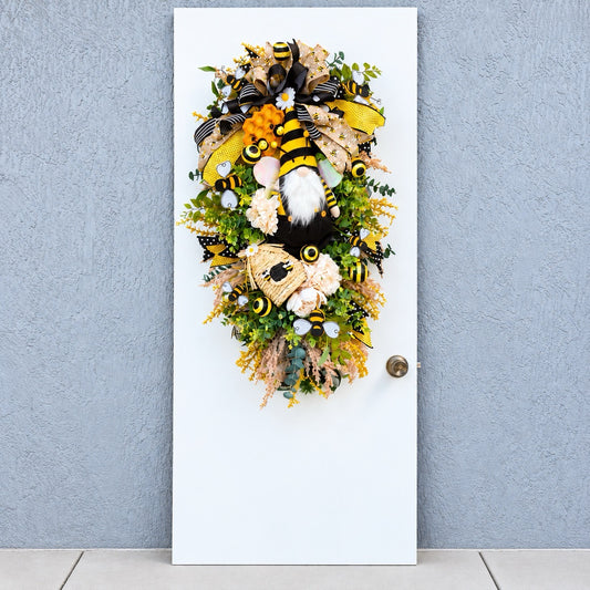 Handmade Queen Bee Manor wreath featuring a bee gnome, woven beehive, yellow and black designer bow, honeycomb accents, florals, bees, and lush greenery.