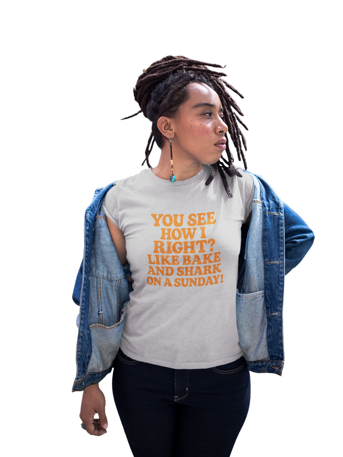 Woman wearing a light pink T-shirt with orange text reading “You see how I right? Like bake and shark on a Sunday!” and an open denim jacket against a white background.