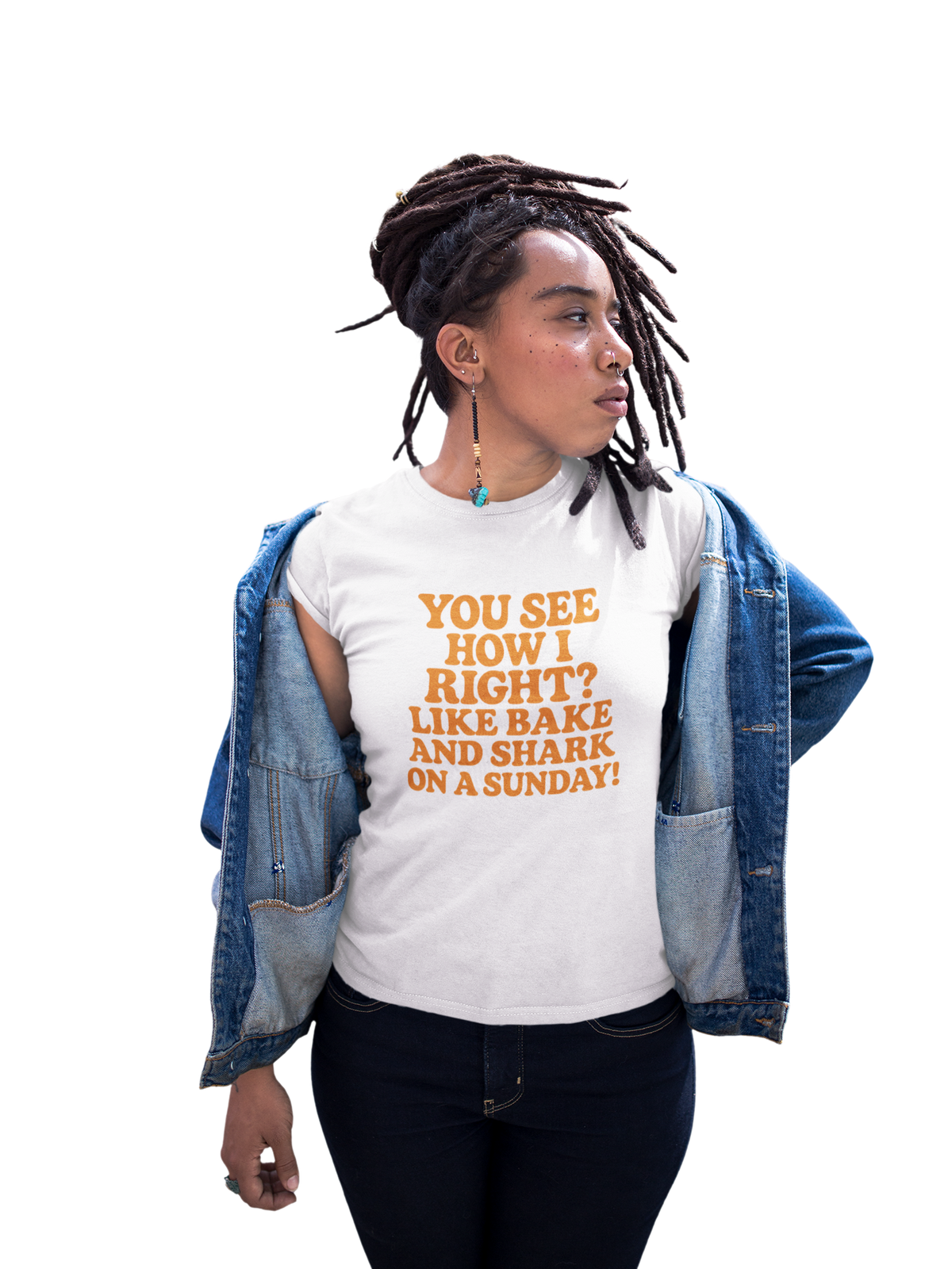 Woman wearing a light pink T-shirt with orange text reading “You see how I right? Like bake and shark on a Sunday!” and an open denim jacket against a white background.
