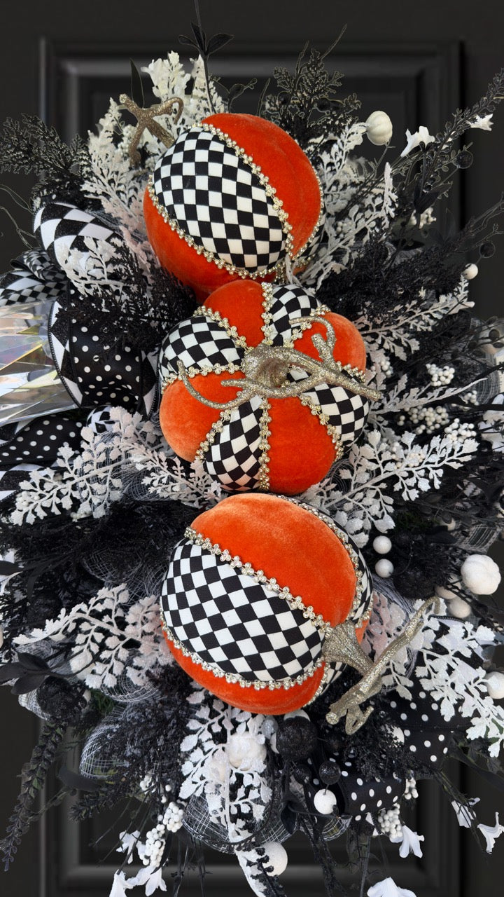 Handmade fall wreath with black‑and‑white checkered pumpkins, gold stems, and black, white, and silver foliage with patterned ribbons on a dark door