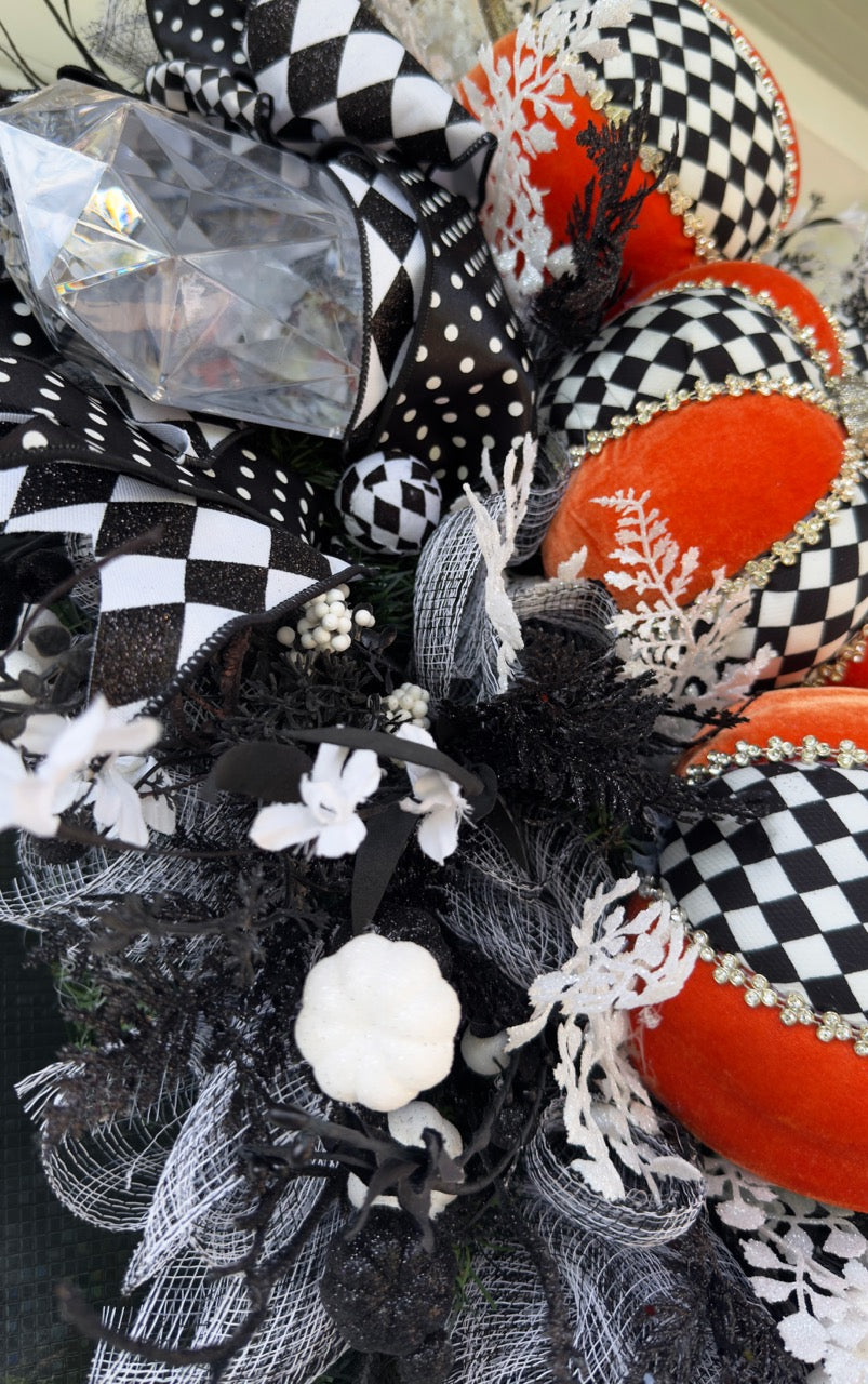 Handmade fall wreath with black‑and‑white checkered pumpkins, gold stems, and black, white, and silver foliage with patterned ribbons on a dark door