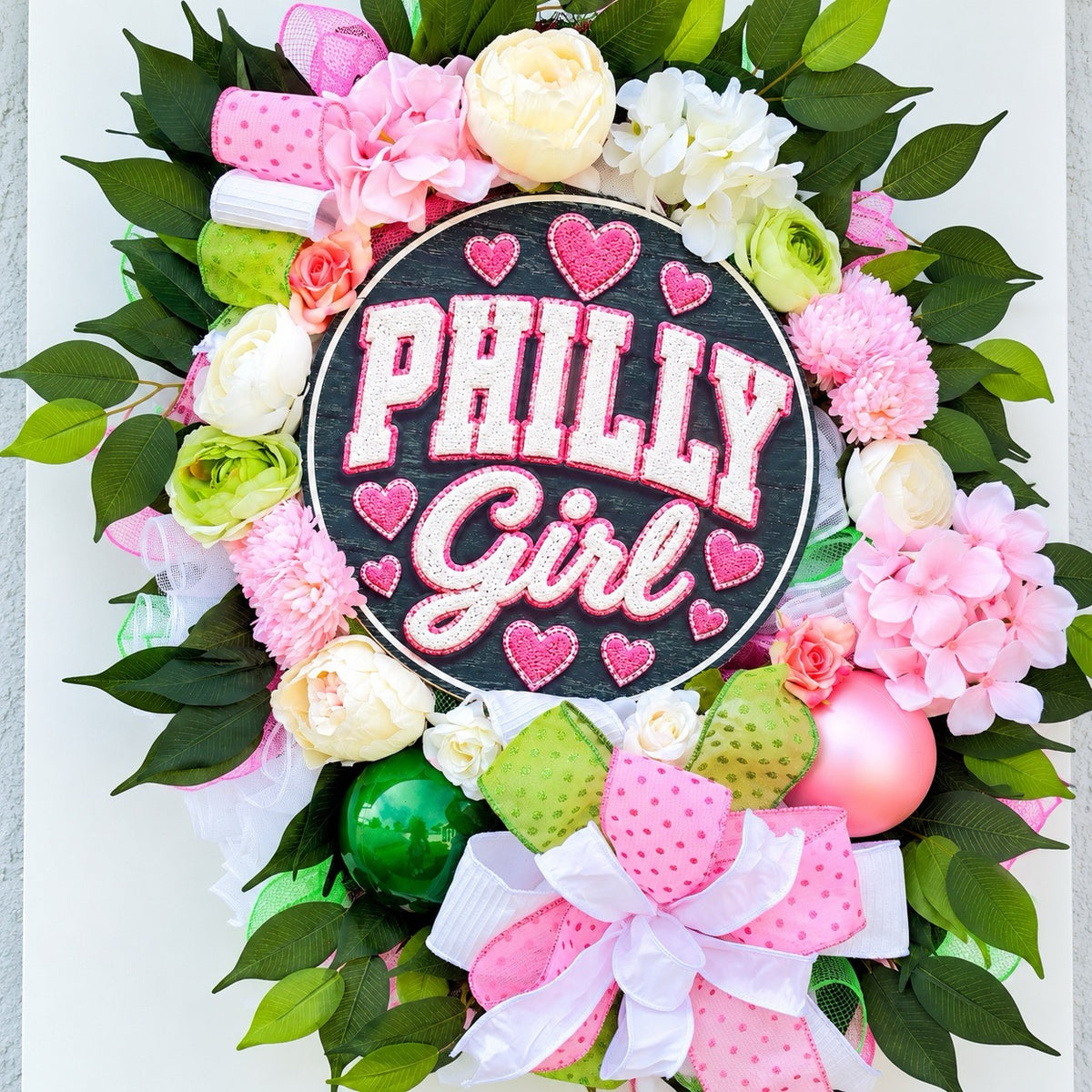 Close-up of black circular Philly Girl wreath sign with sparkling pink lettering and heart accents surrounded by artificial flowers.