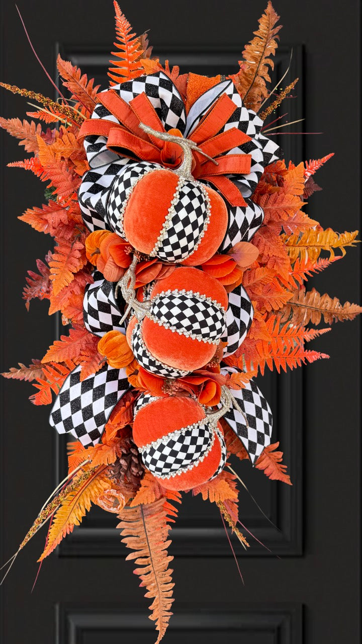 Luxury handmade fall door swag featuring orange velvet pumpkins with black-and-white harlequin accents, designer ribbon bows, crystal trim, gold stems, and vibrant orange autumn ferns on a black front door.