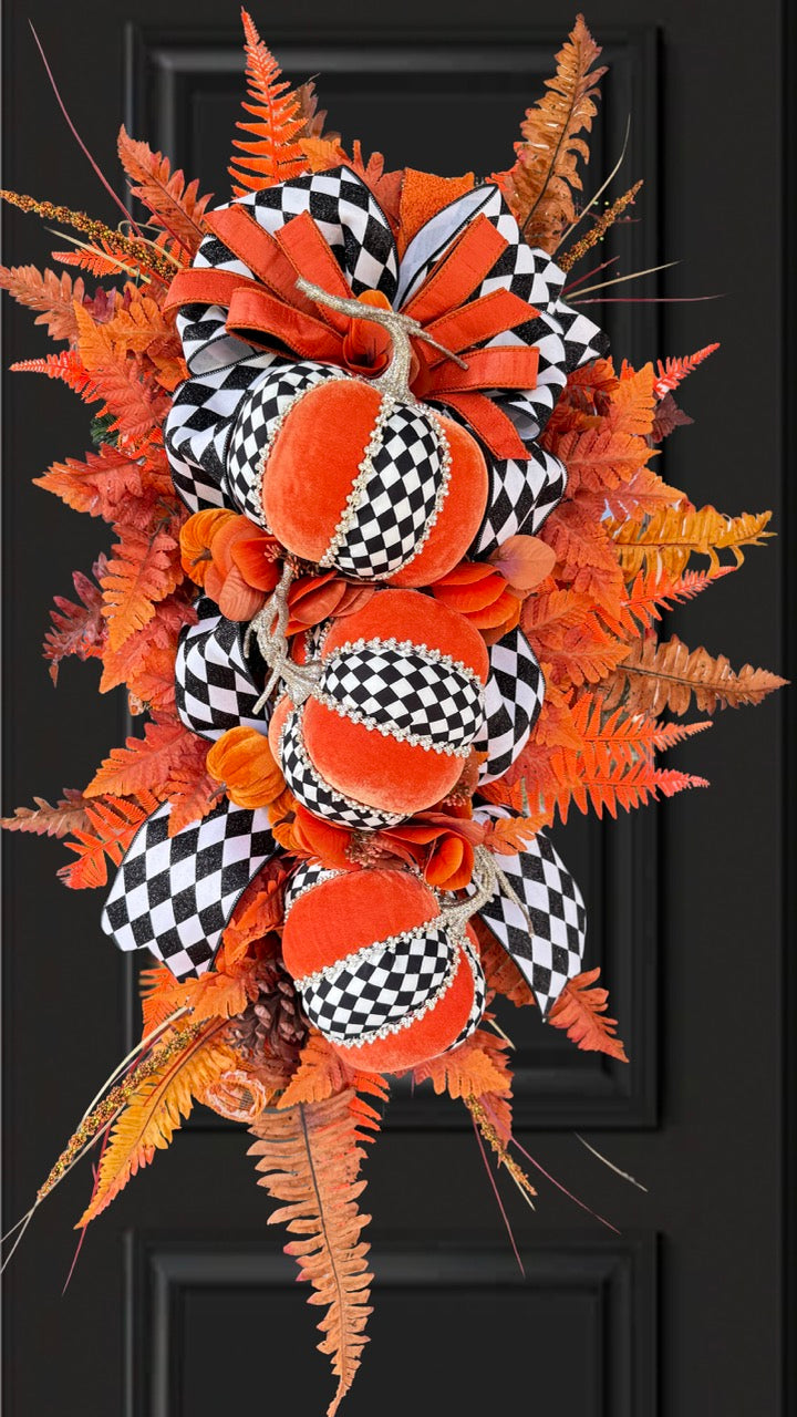Luxury handmade fall door swag featuring orange velvet pumpkins with black-and-white harlequin accents, designer ribbon bows, crystal trim, gold stems, and vibrant orange autumn ferns on a black front door.