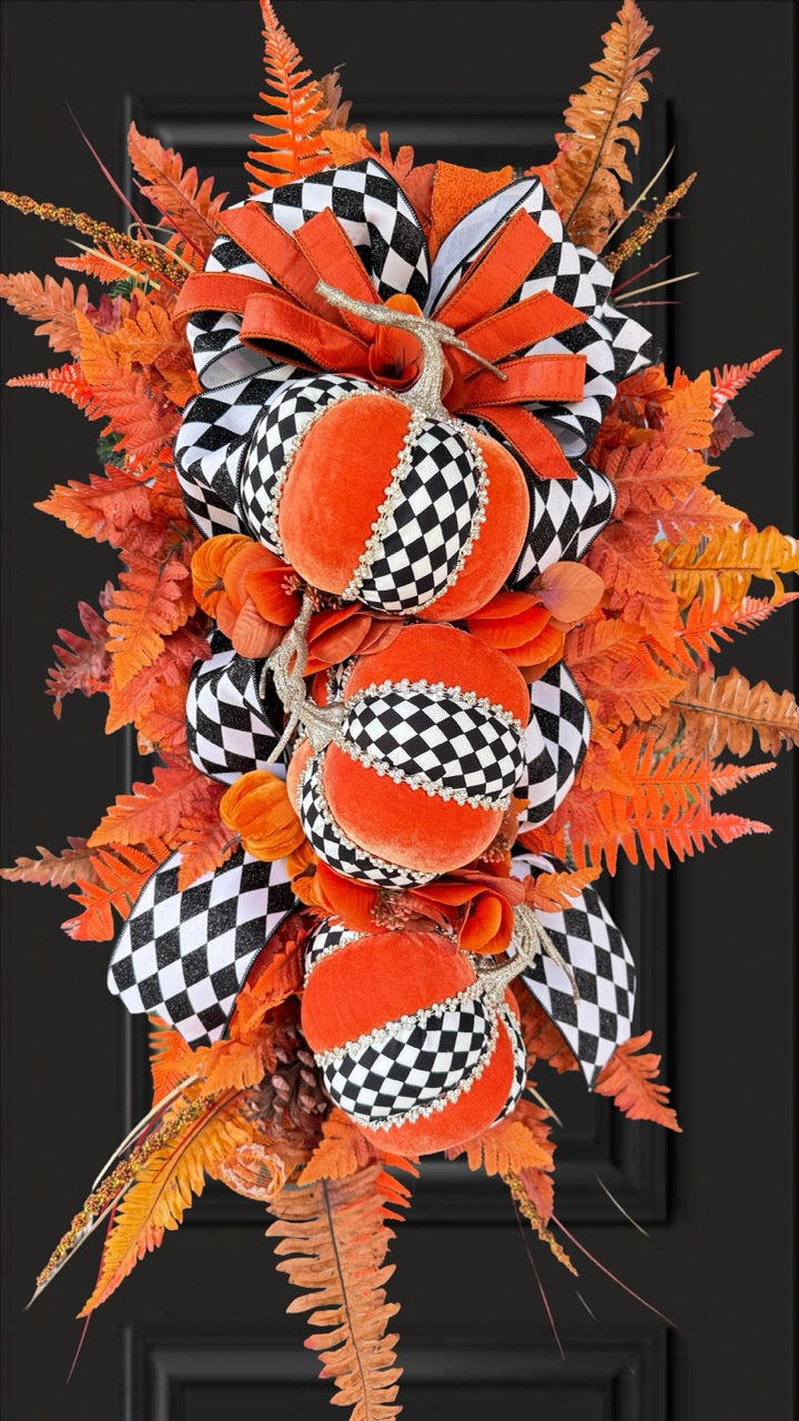 Luxury handmade fall door swag featuring orange velvet pumpkins with black-and-white harlequin accents, designer ribbon bows, crystal trim, gold stems, and vibrant orange autumn ferns on a black front door.