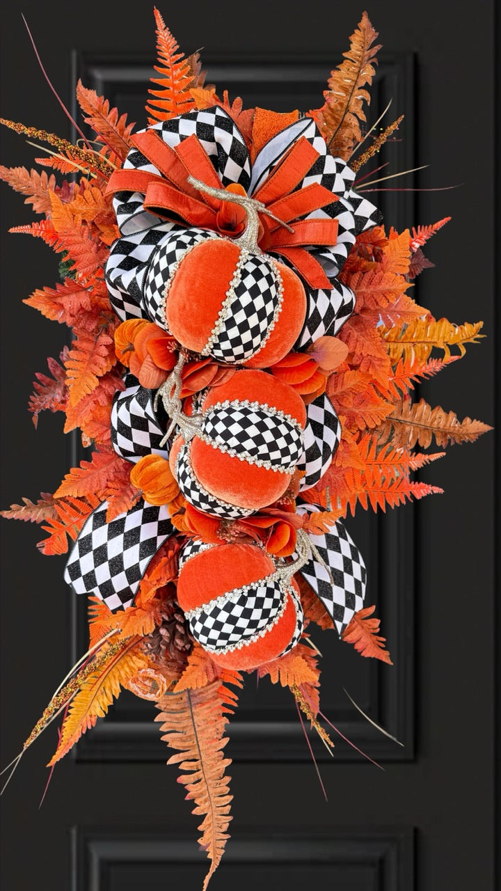 Luxury handmade fall door swag featuring orange velvet pumpkins with black-and-white harlequin accents, designer ribbon bows, crystal trim, gold stems, and vibrant orange autumn ferns on a black front door.