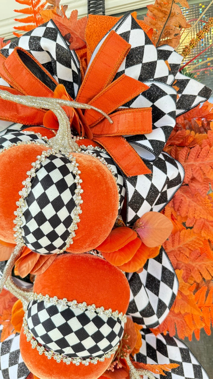 Luxury handmade fall door swag featuring orange velvet pumpkins with black-and-white harlequin accents, designer ribbon bows, crystal trim, gold stems, and vibrant orange autumn ferns on a black front door.