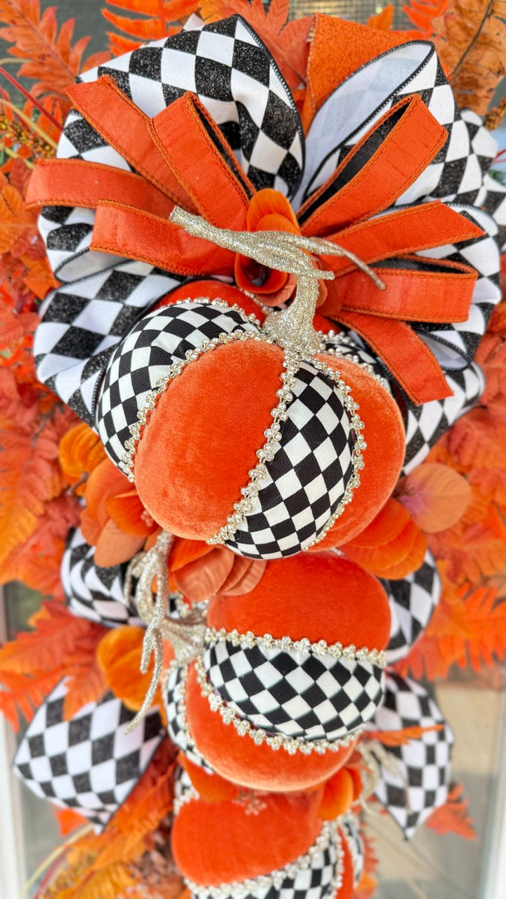 Luxury handmade fall door swag featuring orange velvet pumpkins with black-and-white harlequin accents, designer ribbon bows, crystal trim, gold stems, and vibrant orange autumn ferns on a black front door.