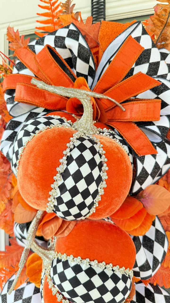 Luxury handmade fall door swag featuring orange velvet pumpkins with black-and-white harlequin accents, designer ribbon bows, crystal trim, gold stems, and vibrant orange autumn ferns on a black front door.