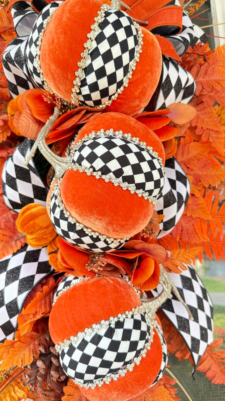 Luxury handmade fall door swag featuring orange velvet pumpkins with black-and-white harlequin accents, designer ribbon bows, crystal trim, gold stems, and vibrant orange autumn ferns on a black front door.