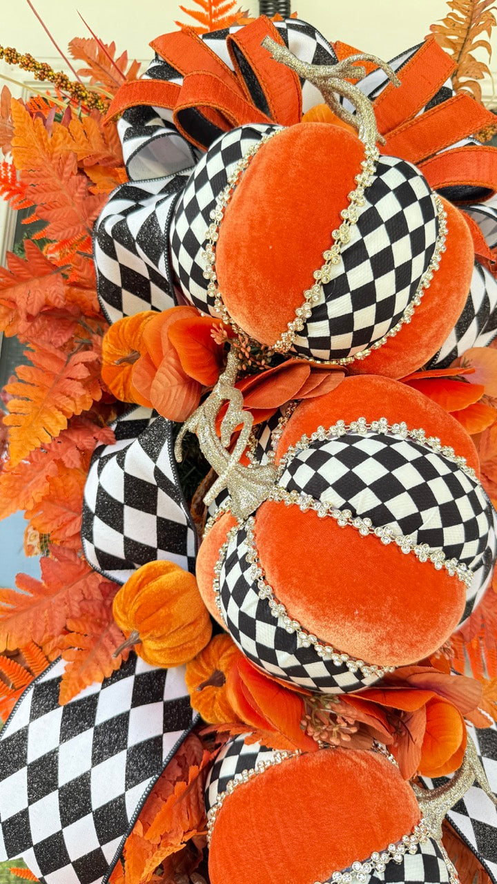 Luxury handmade fall door swag featuring orange velvet pumpkins with black-and-white harlequin accents, designer ribbon bows, crystal trim, gold stems, and vibrant orange autumn ferns on a black front door.