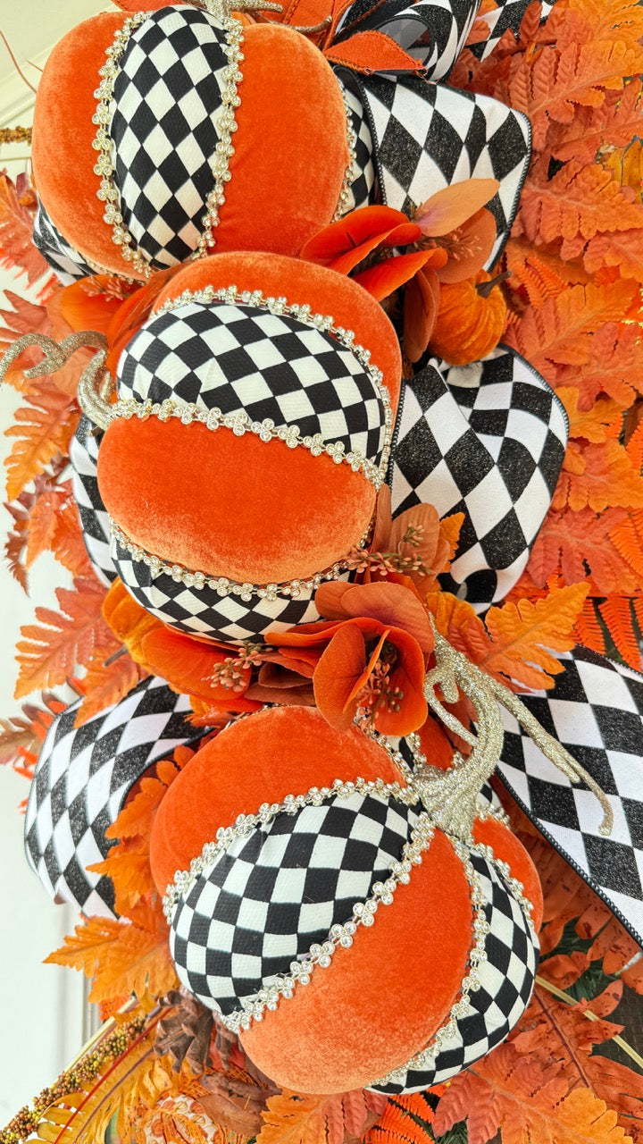 Luxury handmade fall door swag featuring orange velvet pumpkins with black-and-white harlequin accents, designer ribbon bows, crystal trim, gold stems, and vibrant orange autumn ferns on a black front door.