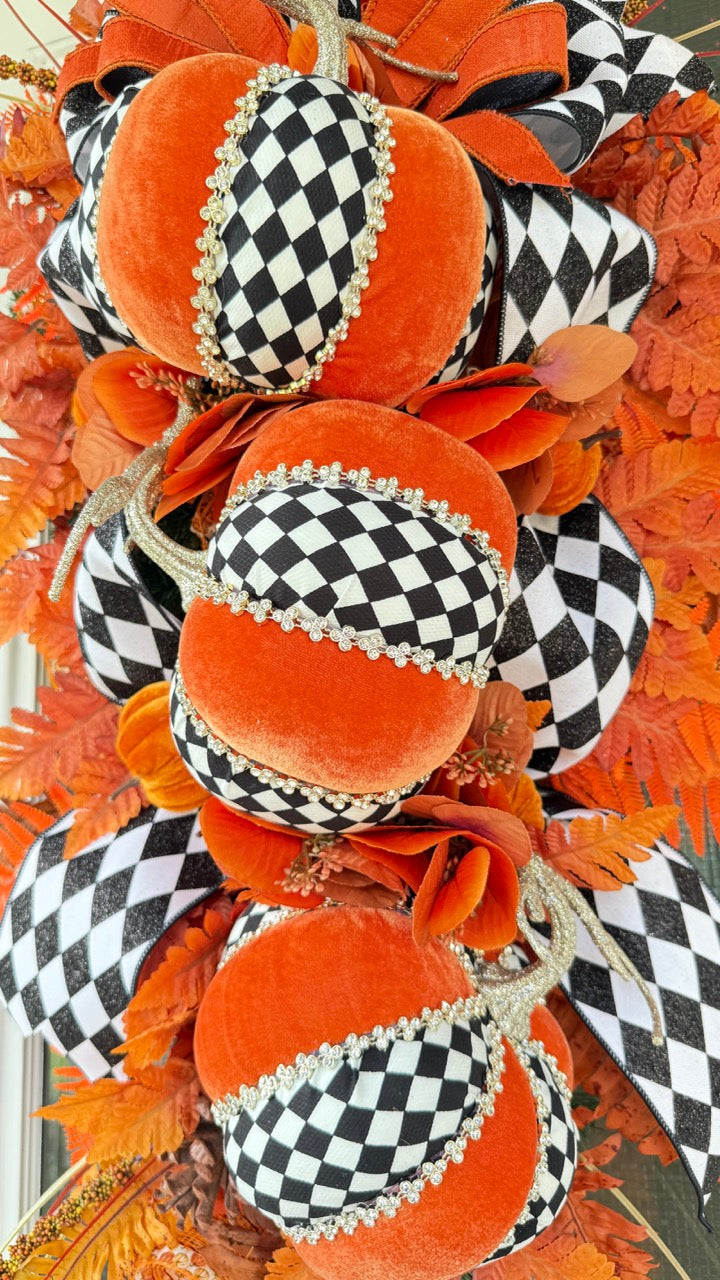 Luxury handmade fall door swag featuring orange velvet pumpkins with black-and-white harlequin accents, designer ribbon bows, crystal trim, gold stems, and vibrant orange autumn ferns on a black front door.