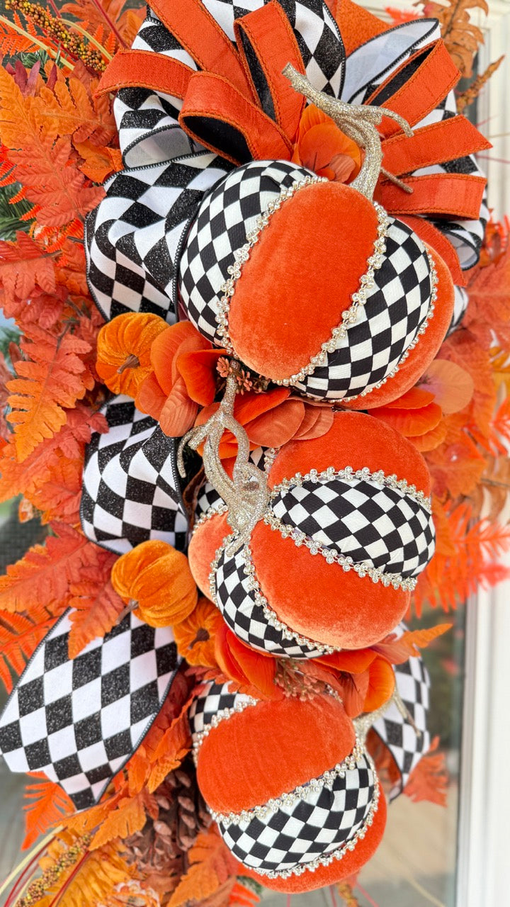 Luxury handmade fall door swag featuring orange velvet pumpkins with black-and-white harlequin accents, designer ribbon bows, crystal trim, gold stems, and vibrant orange autumn ferns on a black front door.