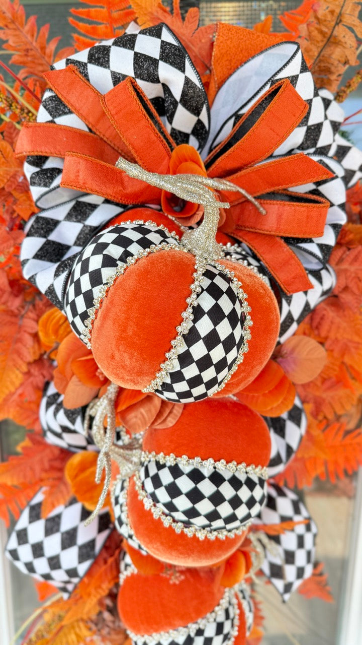 Luxury handmade fall door swag featuring orange velvet pumpkins with black-and-white harlequin accents, designer ribbon bows, crystal trim, gold stems, and vibrant orange autumn ferns on a black front door.