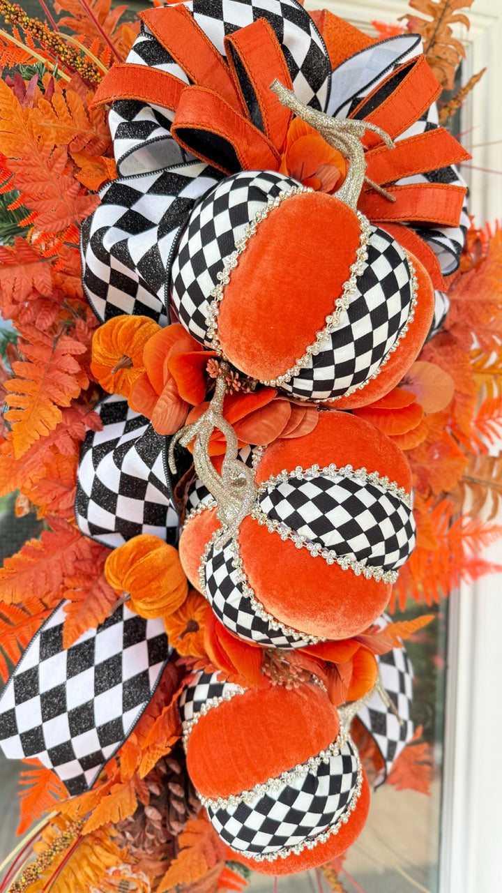 Luxury handmade fall door swag featuring orange velvet pumpkins with black-and-white harlequin accents, designer ribbon bows, crystal trim, gold stems, and vibrant orange autumn ferns on a black front door.
