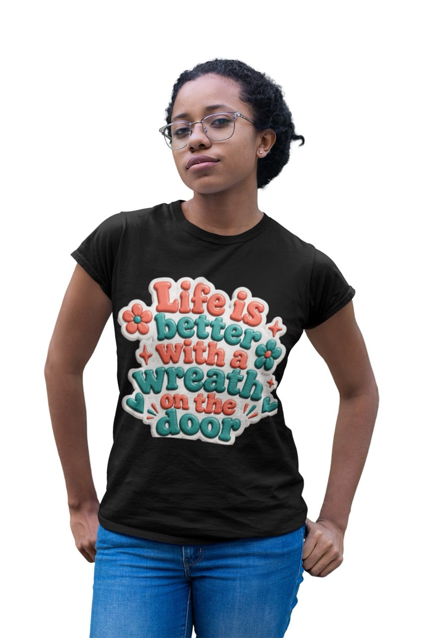 Graphic t-shirt featuring the phrase “Life Is Better With a Wreath on the Door” in playful dimensional lettering with decorative flowers and accent elements.