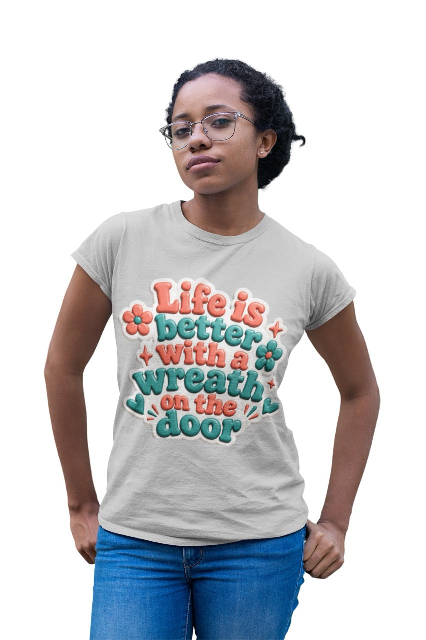 Graphic t-shirt featuring the phrase “Life Is Better With a Wreath on the Door” in playful dimensional lettering with decorative flowers and accent elements.