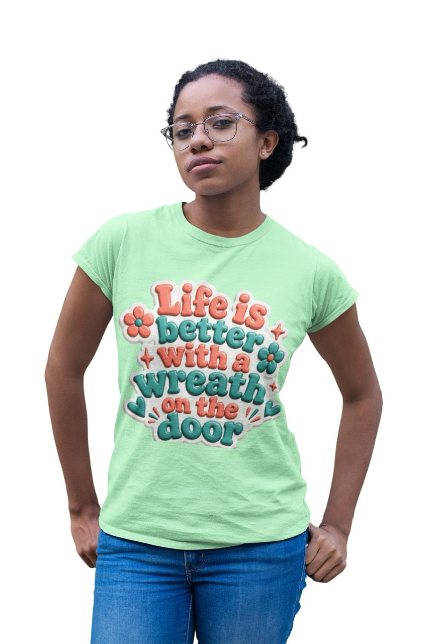 Graphic t-shirt featuring the phrase “Life Is Better With a Wreath on the Door” in playful dimensional lettering with decorative flowers and accent elements.