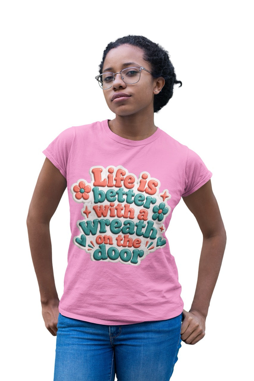 Graphic t-shirt featuring the phrase “Life Is Better With a Wreath on the Door” in playful dimensional lettering with decorative flowers and accent elements.