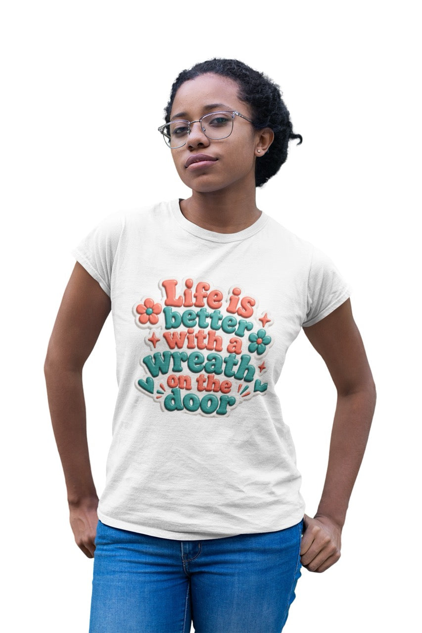 Graphic t-shirt featuring the phrase “Life Is Better With a Wreath on the Door” in playful dimensional lettering with decorative flowers and accent elements.