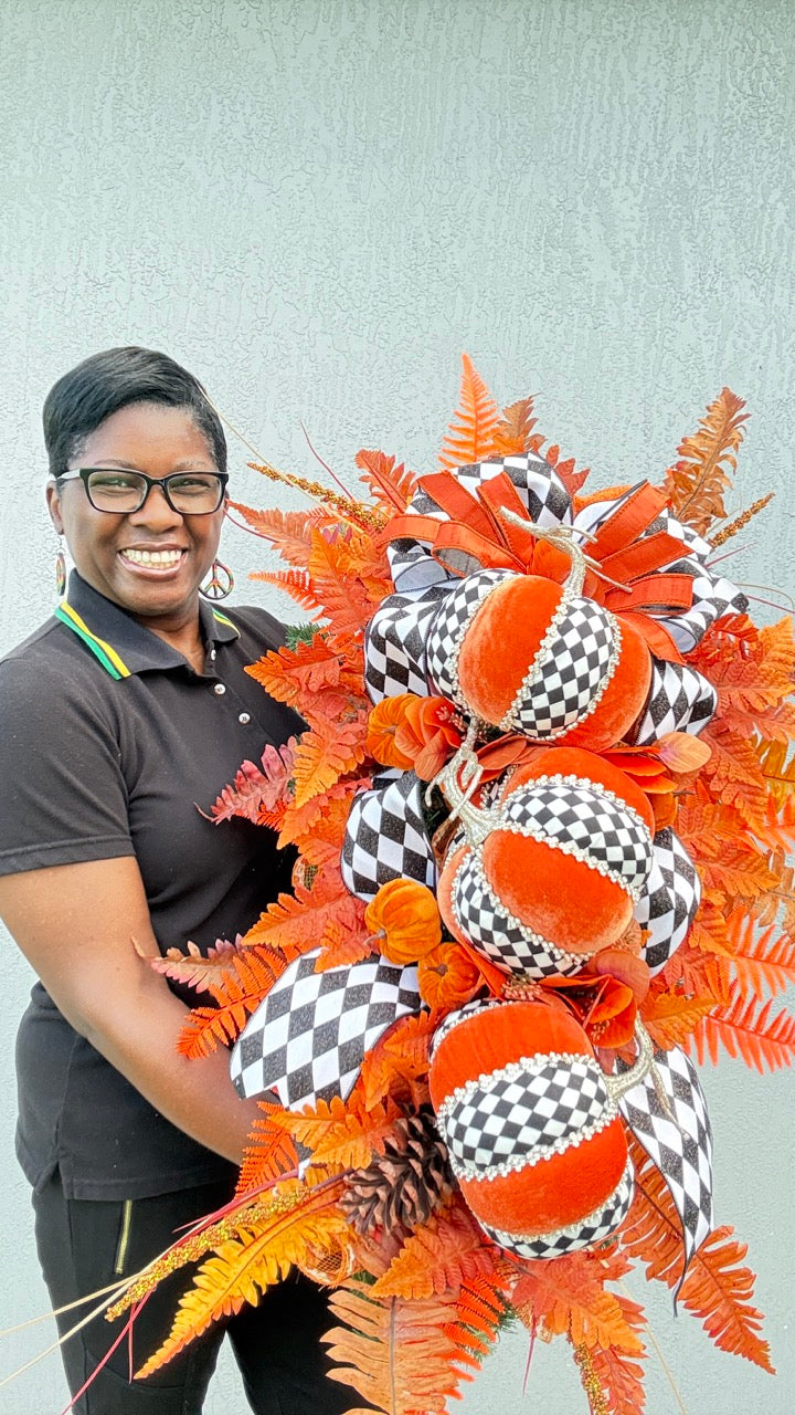 Luxury handmade fall door swag featuring orange velvet pumpkins with black-and-white harlequin accents, designer ribbon bows, crystal trim, gold stems, and vibrant orange autumn ferns on a black front door.