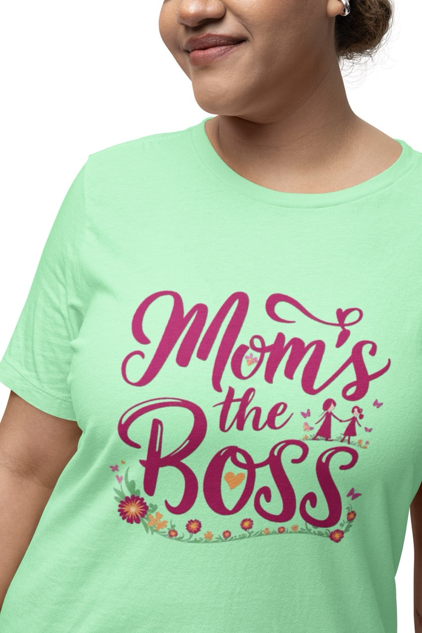 Woman wearing a green T-shirt with bold pink script reading “Mom’s the Boss,” decorated with flowers, hearts, and a small illustration of a mother and child.