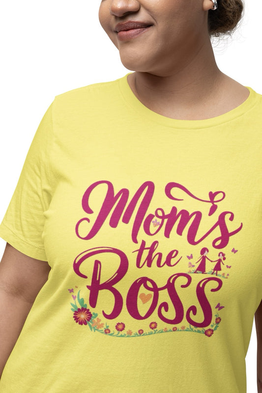 Woman wearing a yellow T-shirt with bold pink script reading “Mom’s the Boss,” decorated with flowers, hearts, and a small illustration of a mother and child.