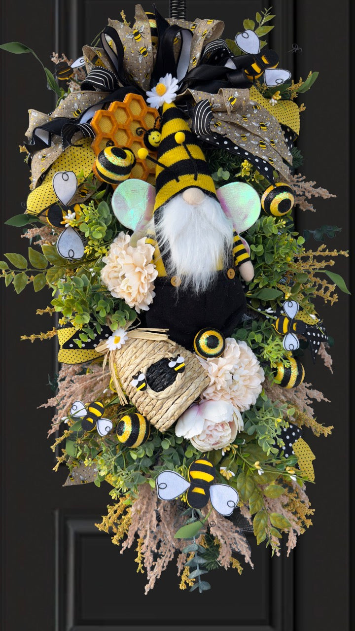 Luxury bee-themed door swag featuring a whimsical bee gnome, honeycomb decoration, beehive accent, premium black and yellow ribbons, lush greenery, cream florals, and sparkling bee embellishments, perfect for spring and summer front door décor.