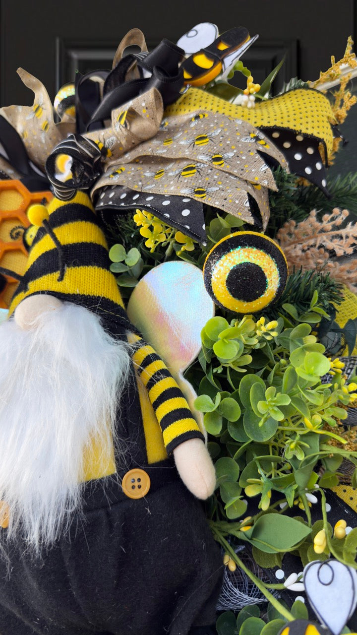 Luxury bee-themed door swag featuring a whimsical bee gnome, honeycomb decoration, beehive accent, premium black and yellow ribbons, lush greenery, cream florals, and sparkling bee embellishments, perfect for spring and summer front door décor.
