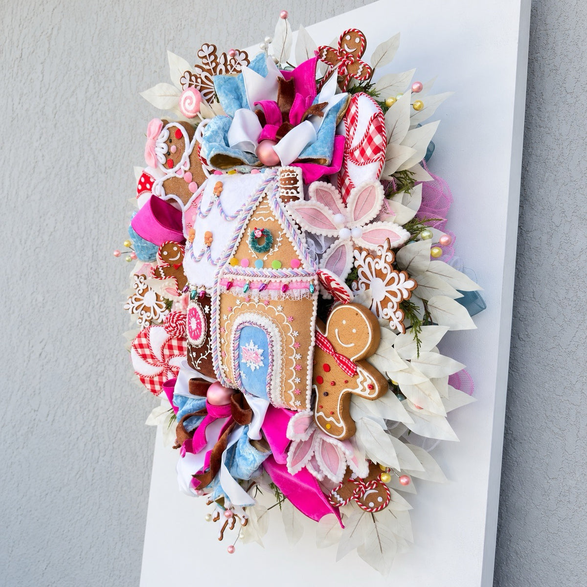 Sugarplum Gingerbread Cottage Christmas Wreath with pastel gingerbread house, pink and blue bows, poinsettias and Norfolk fir greenery.