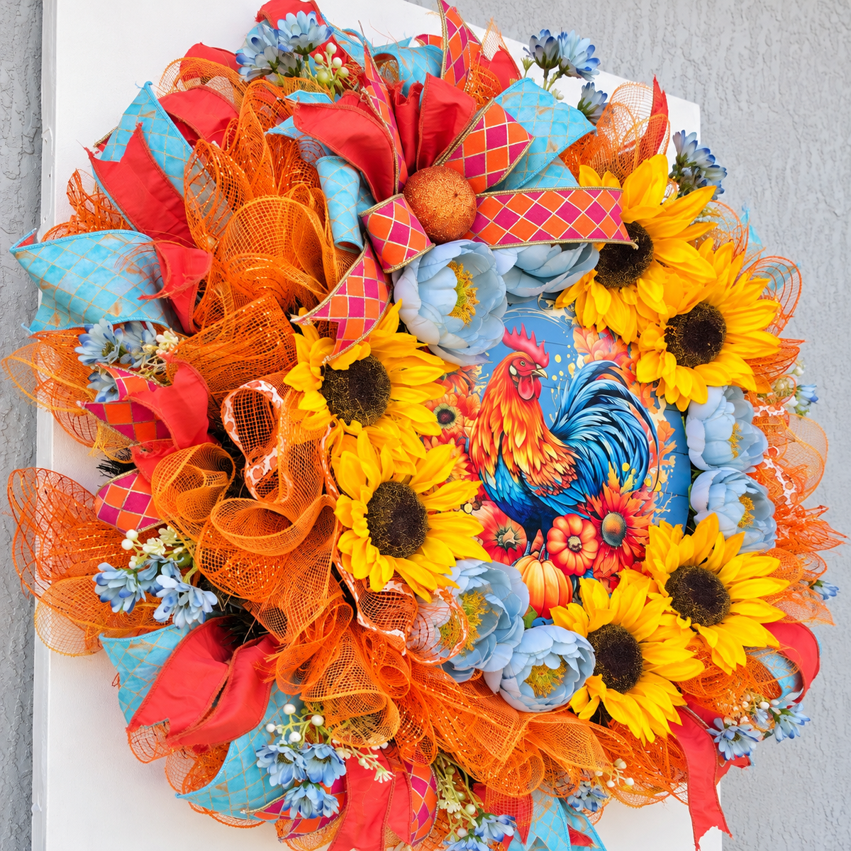 Handmade fall rooster wreath featuring vibrant yellow sunflowers, dusty blue flowers, orange deco mesh, coral and turquoise ribbons, and a colorful rooster centerpiece. A full farmhouse-style autumn wreath displayed on a white front door.