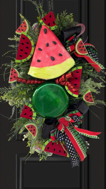 Handmade watermelon summer swag wreath featuring oversized watermelon slice accents, glitter watermelon embellishments, designer black and red ribbon bows, lush greenery, and festive farmhouse décor displayed on a black front door.