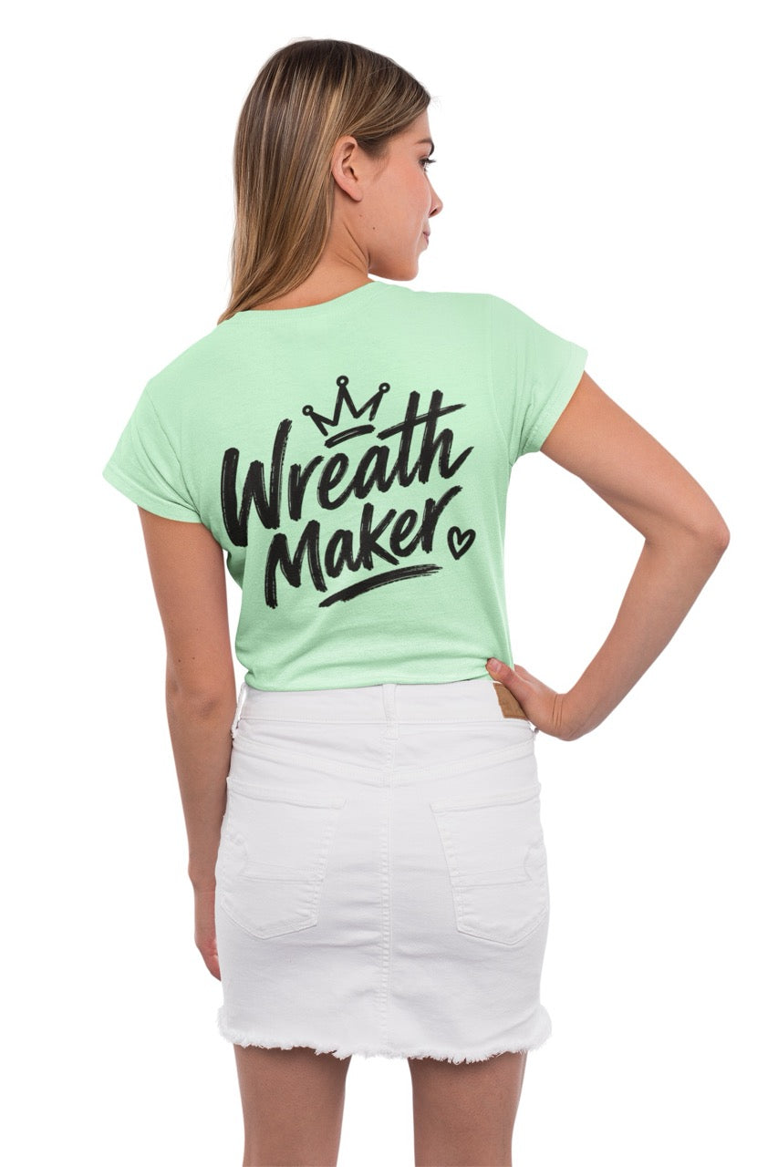 Back view of a t-shirt displaying the words “Wreath Maker” in large brush-style lettering with a crown above and a small heart beside the design. The shirt is available in various colors.