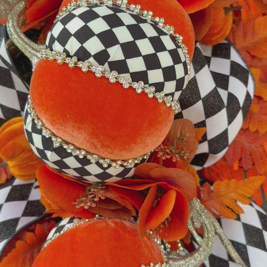 Luxury handmade fall door swag featuring orange velvet pumpkins with black-and-white harlequin accents, designer ribbon bows, crystal trim, gold stems, and vibrant orange autumn ferns on a black front door.