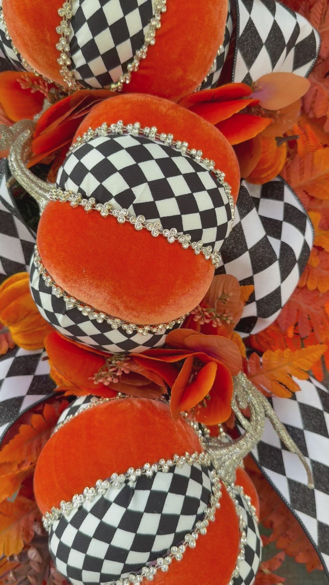 Luxury handmade fall door swag featuring orange velvet pumpkins with black-and-white harlequin accents, designer ribbon bows, crystal trim, gold stems, and vibrant orange autumn ferns on a black front door.