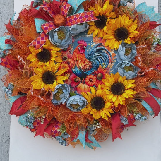 Handmade fall rooster wreath featuring vibrant yellow sunflowers, dusty blue flowers, orange deco mesh, coral and turquoise ribbons, and a colorful rooster centerpiece. A full farmhouse-style autumn wreath displayed on a white front door.