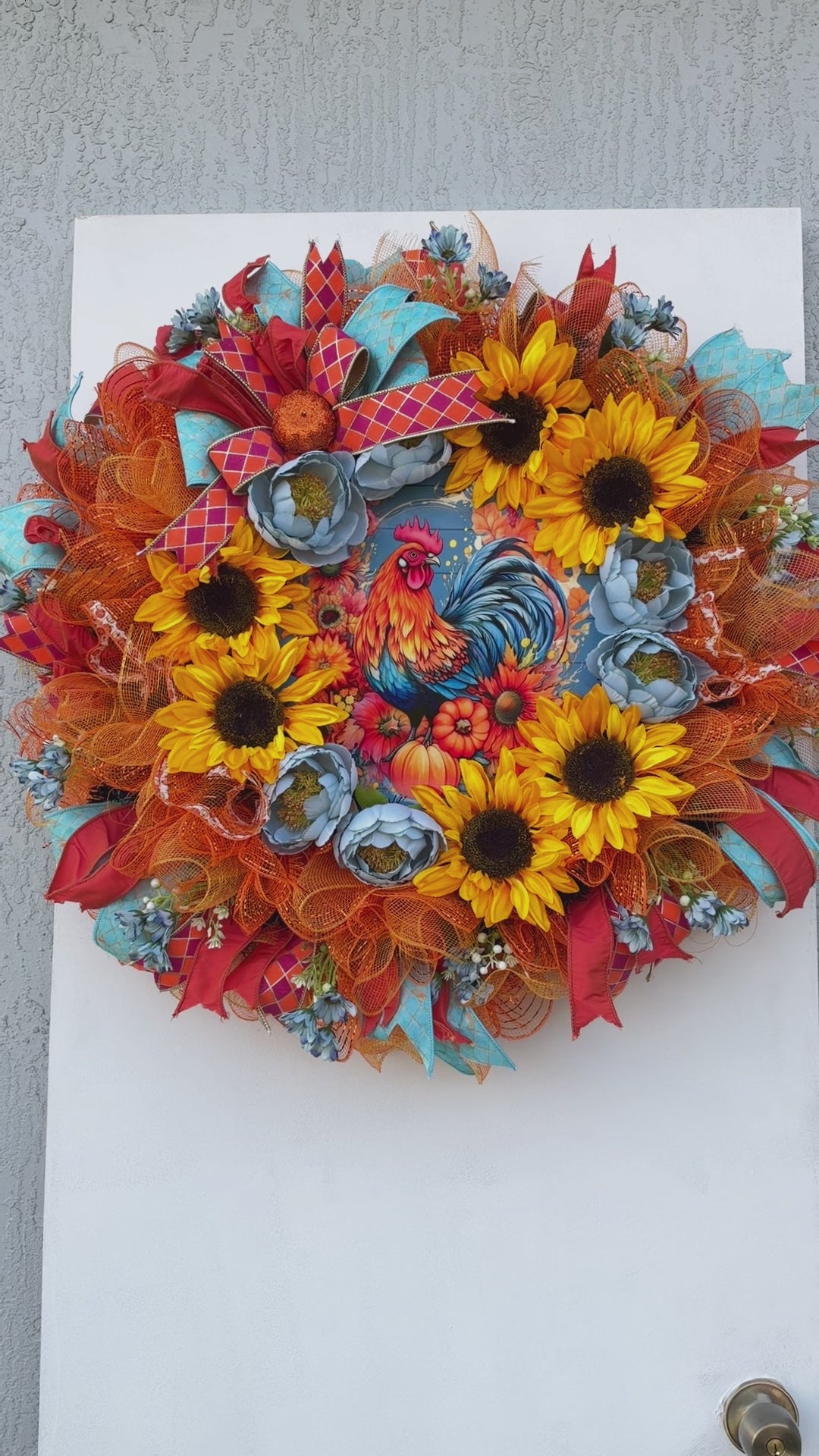 Handmade fall rooster wreath featuring vibrant yellow sunflowers, dusty blue flowers, orange deco mesh, coral and turquoise ribbons, and a colorful rooster centerpiece. A full farmhouse-style autumn wreath displayed on a white front door.
