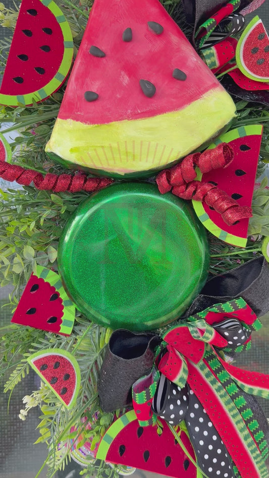 Handmade watermelon summer swag wreath featuring oversized watermelon slice accents, glitter watermelon embellishments, designer black and red ribbon bows, lush greenery, and festive farmhouse décor displayed on a black front door.